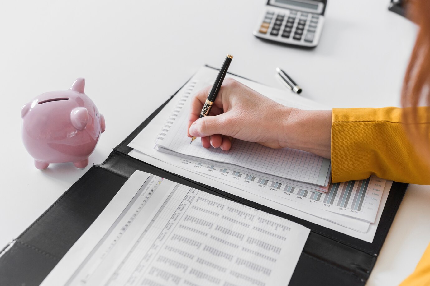 person with a pen writes in a notebook, surrounded by financial documents